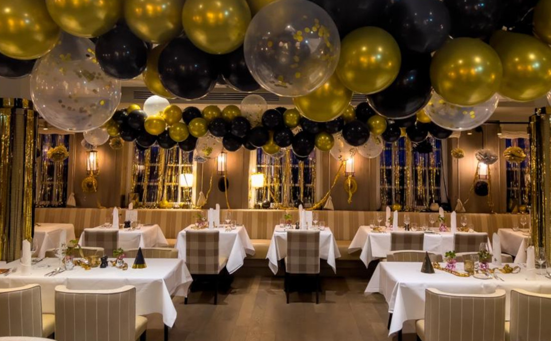 Elegantes Restaurant mit festlicher Dekoration: goldene, schwarze und transparente Luftballons über weiß gedeckten Tischen.