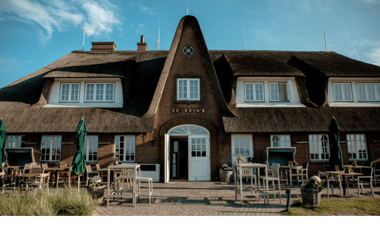 Das Landhaus Severin*s Morsum Kliff mit Reetdach, Terrasse und Gartenmöbeln vor blauem Himmel.
