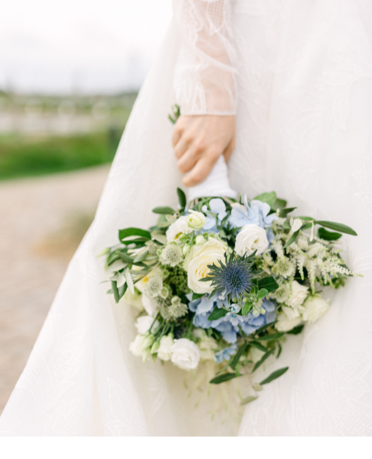 Braut hält Blumenstrauß mit weißen und blauen Blumen vor grüner Landschaft.