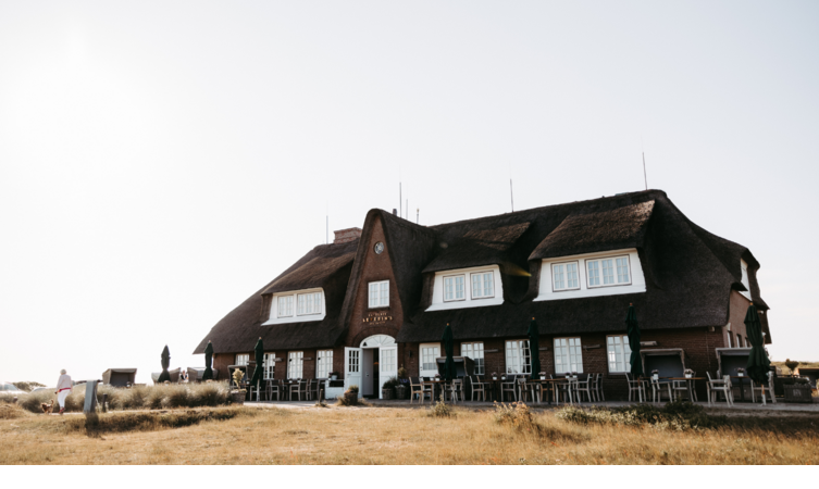 Reetgedecktes Hotelgebäude mit Terrasse, umgeben von Wiese, unter klarem Himmel.