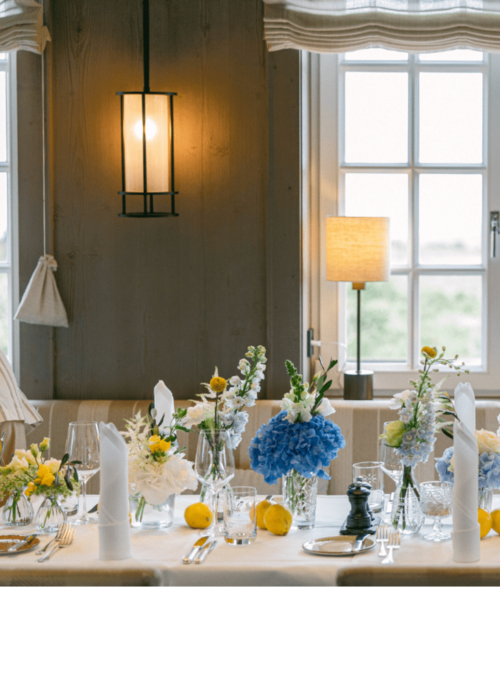 Eleganter, gedeckter Tisch mit blauen und gelben Blumenarrangements vor einem großen Fenster im Landhaus Severin*s Morsum Kliff.