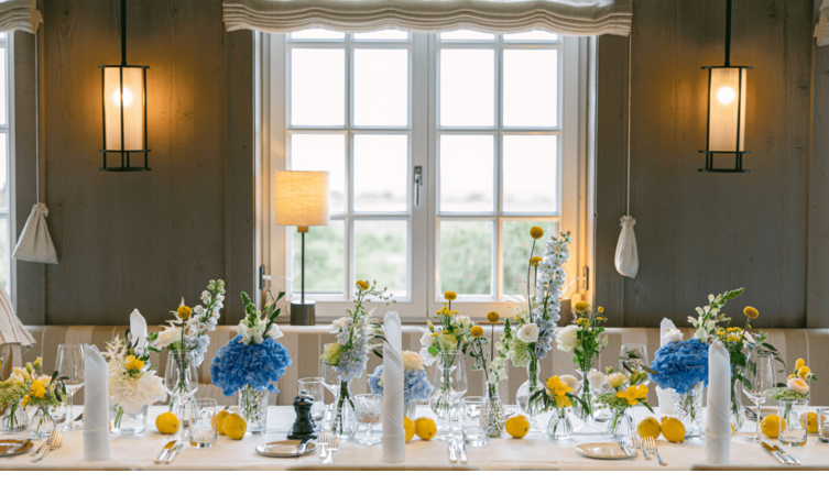 Eleganter, gedeckter Tisch mit blauen und gelben Blumenarrangements vor einem großen Fenster im Landhaus Severin*s Morsum Kliff.