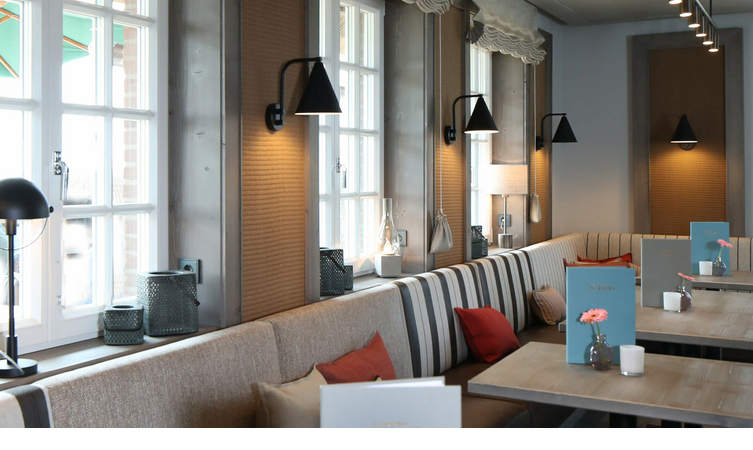 Gemütliche Atmosphäre in der Kamin-Bar im Landhaus Severin*s auf Sylt Gemütliches Restaurant mit gestreiften Sitzbänken, Holztischen, Blumenvasen und großen Fenstern im Hotel Landhaus Severin*s.