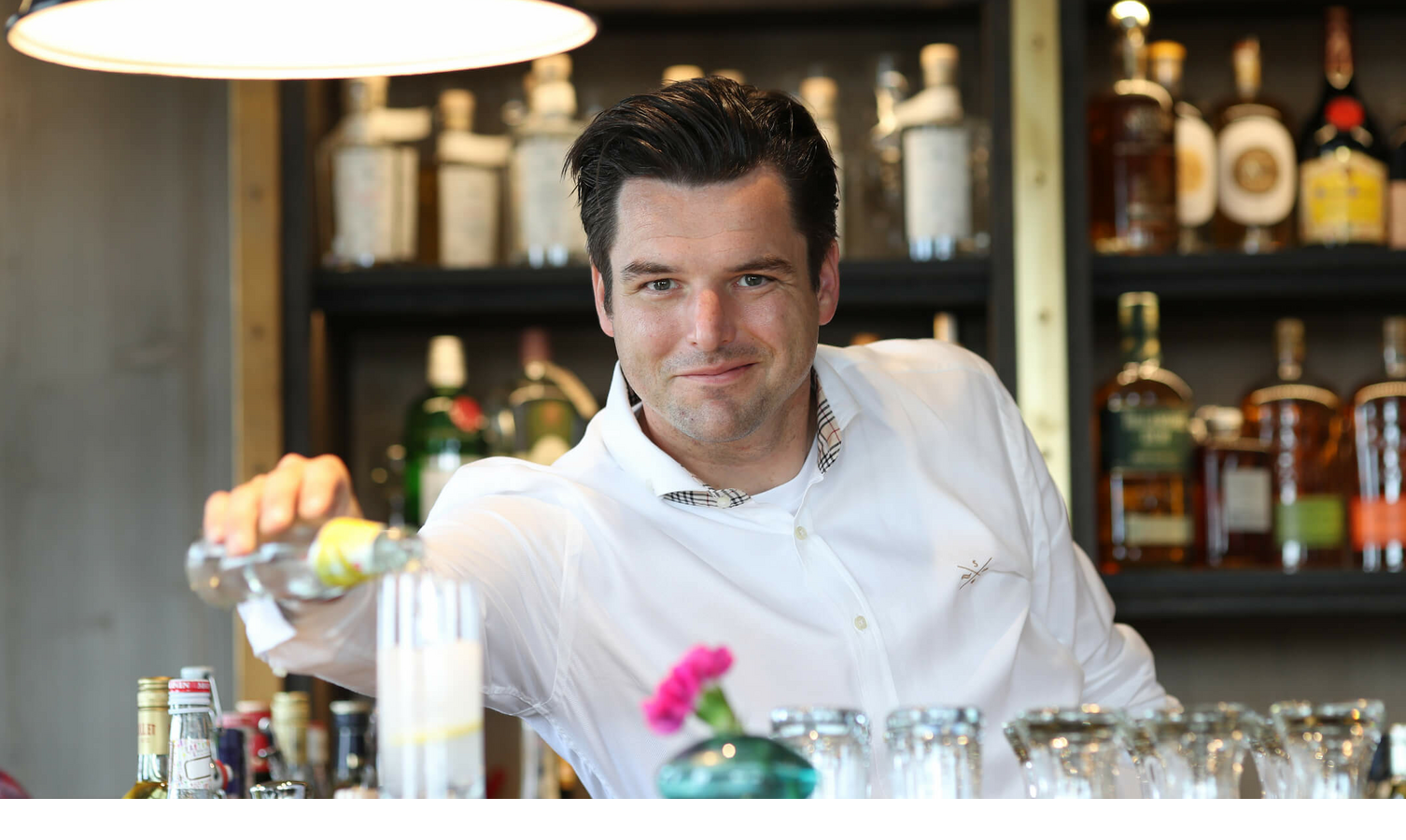 Barkeeper in weißem Hemd schenkt Getränk an stilvoller Hotelbar ein, im Hintergrund Flaschenregal.