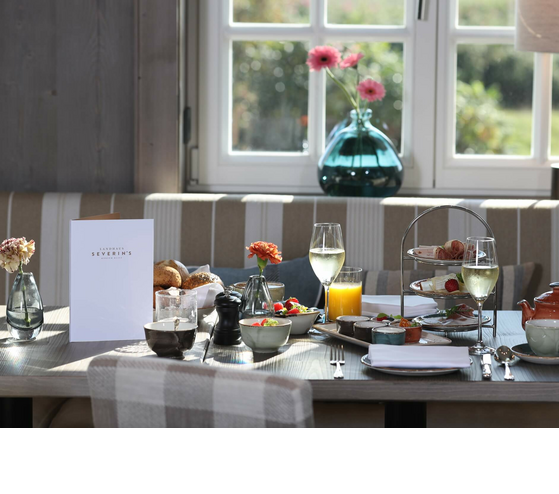 Frühstückstisch im Landhaus Severin*s mit Gebäck, Getränken und Blumen vor einem Fenster.