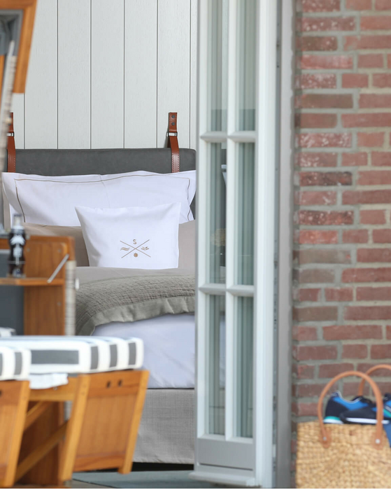 Gemütliche Hotelterrasse mit Strandkorb, Blick ins helle Zimmer mit Bett, Korbtasche im Vordergrund.