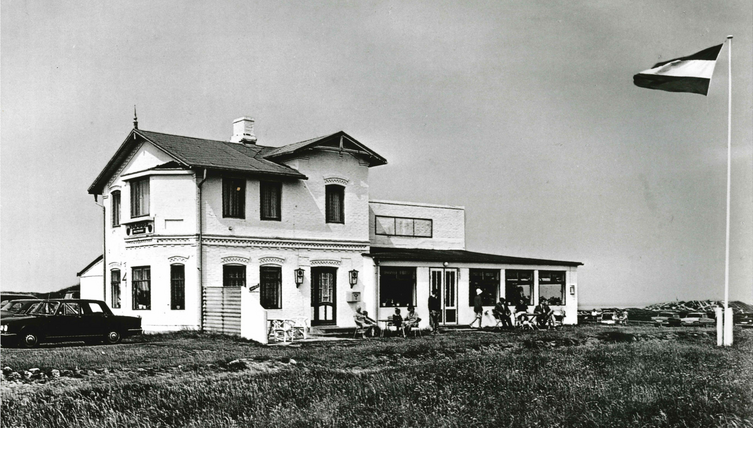 Historisches Hotel am Strand mit Terrasse und Gästen, daneben weht eine Flagge.