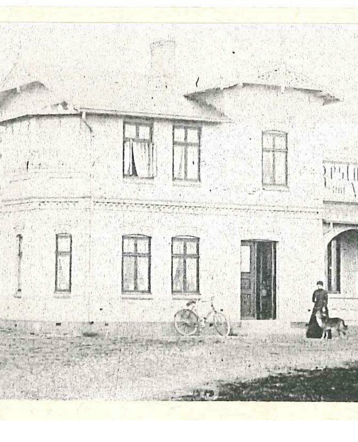 Historisches Hotelgebäude mit Veranda, Fahrrad und Person mit Hund im Vordergrund.