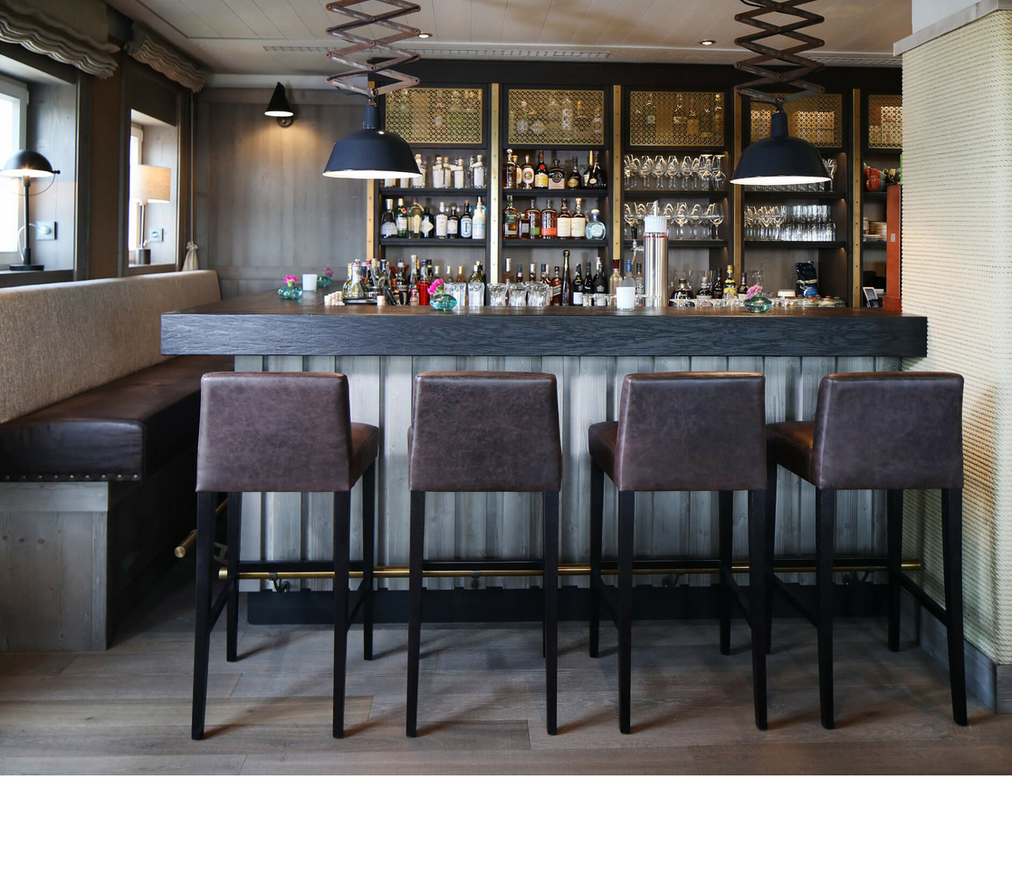 Gemütliche Hotelbar mit Holztheke, vier braunen Barhockern und gut bestücktem Regal im Hintergrund.