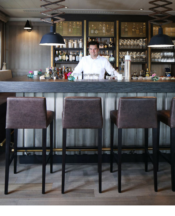 Gemütliche Hotelbar mit Theke, vier Barhockern und einem Barkeeper im Landhaus Severin*s Morsum Kliff auf Sylt.