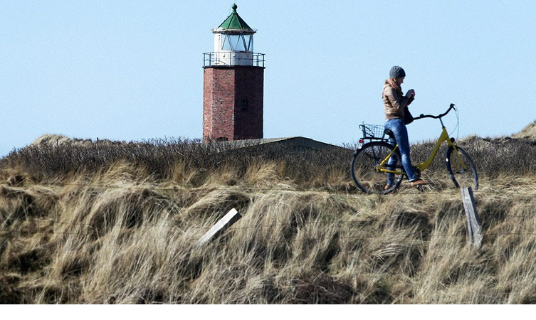 Person auf gelbem Fahrrad vor einem roten Backstein-Leuchtturm in einer grasbewachsenen Dünenlandschaft.