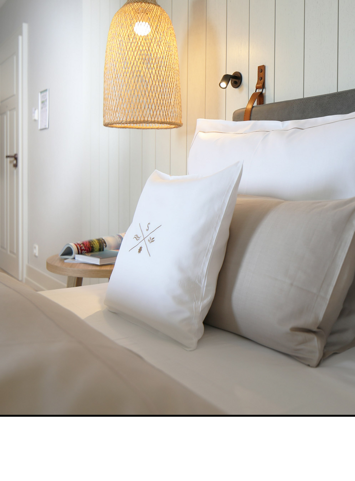 Gemütliches Hotelzimmer mit elegantem Bett, weißen Kissen, hölzernem Nachttisch und geflochtener Hängelampe.