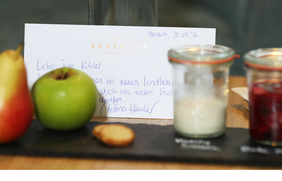 Karte mit Willkommensnachricht, Apfel, Birne, Keks und zwei Gläser mit Joghurt und Marmelade auf einem Tisch.