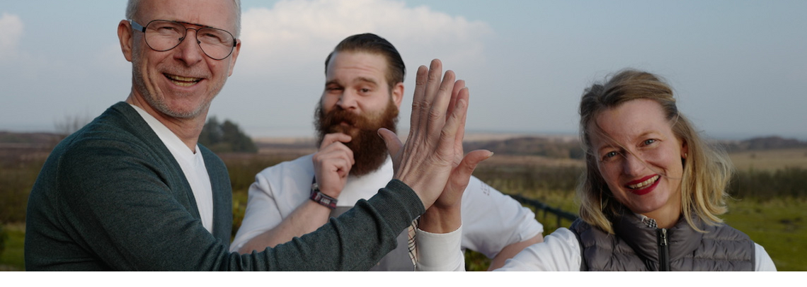 Drei Menschen lächeln in die Kamera, zwei geben sich ein High-Five, im Hintergrund eine Landschaft.